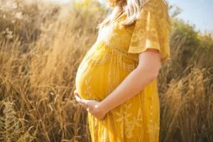 pregnant woman wearing yellow floral dress standing while touching her tummy and facing her right side near brown field during daytime.Photo by Anna Hecker